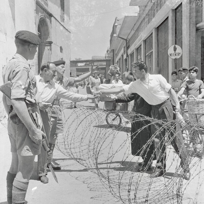 In this historical photograph of Nicosia's Hermes Street, Cypriots make cash transactions across barbed wire barrier while a British soldier stands guard.