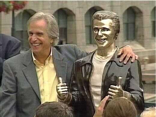 Henry Winkler posing for a picture beside a bronze statue of Arthur Fonzarelli, also known as fonzie, from tv show happy days.